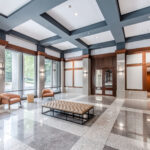 Elegant office lobby featuring coffered ceiling, marble floors, wood paneling, leather seating, and floor-to-ceiling windows with natural light