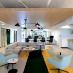 Modern open workspace featuring grey sectional sofa, light blue accent chairs, geometric floor patterns in green and yellow, wood ceiling detail, and black pendant lighting