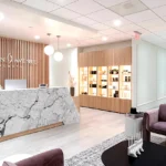 Sophisticated reception area with marble desk, wooden slat wall feature, illuminated product display, mauve accent chairs, and modern lighting in Dallas medical facility