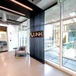 Modern office entrance featuring the LINK logo on black vertical slats, glass walls, outdoor terrace access, and contemporary lounge seating