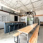 Industrial taproom featuring wooden communal tables, metal bar stools, decorative tile wall, string lighting, and a custom spider decoration suspended from the exposed ceiling