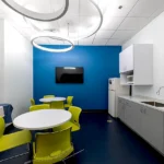Modern break room featuring vibrant blue accent wall, lime green chairs, white round tables, circular pendant lights, and full kitchen amenities