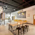 Tasting area with illuminated Schoolhouse Brewing logo sign, industrial brewing tanks, live-edge wood table, and 'Principal's Office' door. School-themed chalkboard art and pendant lighting.