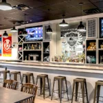 Sports bar featuring custom trophy display shelving, Schoolhouse logo, metal bar stools, dining tables, multiple TVs, and vintage sports memorabilia in Atlanta venue