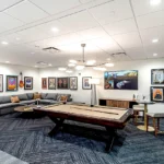 Modern game room featuring a pool table, sectional gray couch, contemporary light fixture, and music-themed wall art collection with a large flat-screen TV displaying surrealist art