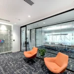 Contemporary reception area at Alliant Health Solutions featuring glass display shelving, coral lounge chairs, glass partition walls, and textured carpeting by Cornerstone Contracting
