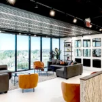 Modern lounge area at Lucid Private Offices Atlanta featuring panoramic windows, custom shelving, grey and orange seating, and textured reception desk by Cornerstone Contracting